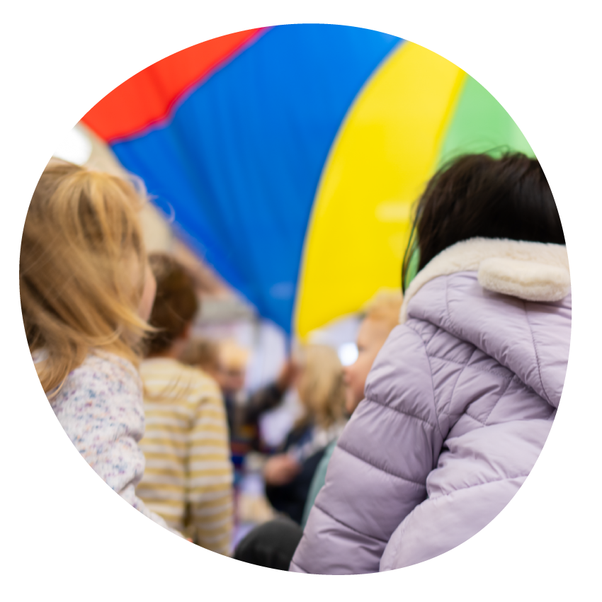 Image: Young children look upwards at a brightly coloured piece of fabric held high in a play activity.