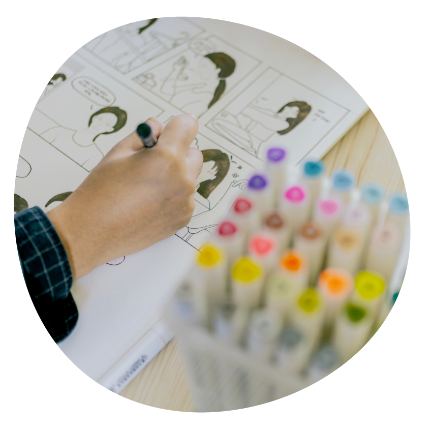 Image: Close-up of a hand drawing a comic strip. Coloured textas sit next to their notebook.