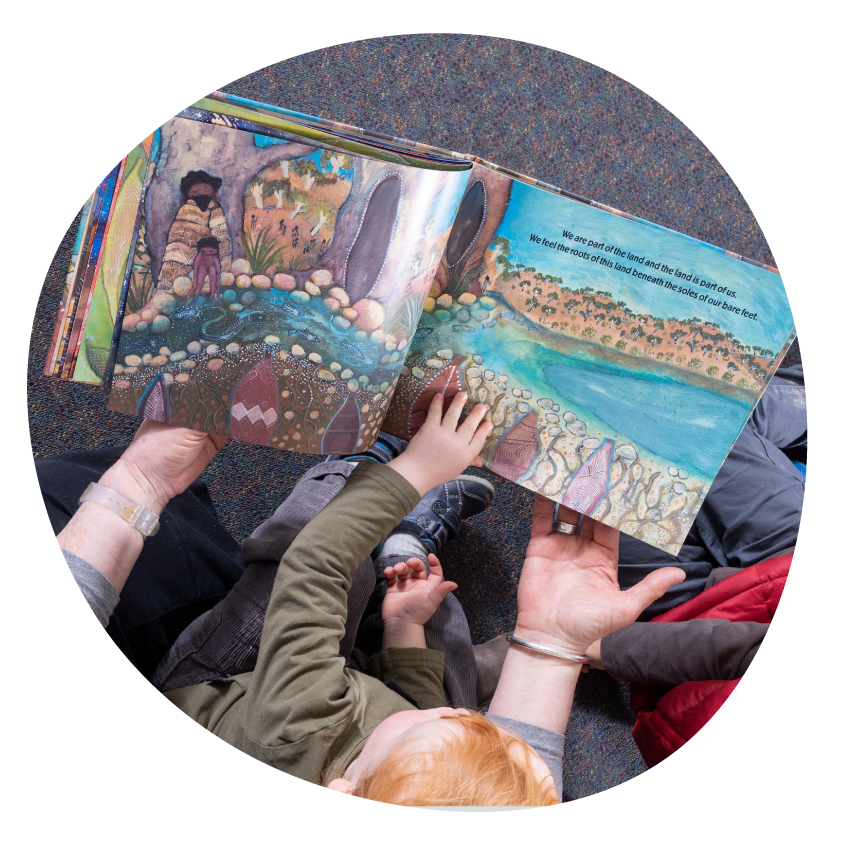 Image: Top-down view of an adult and two children seated on the floor together reading from a picture book featuring Aboriginal artwork.