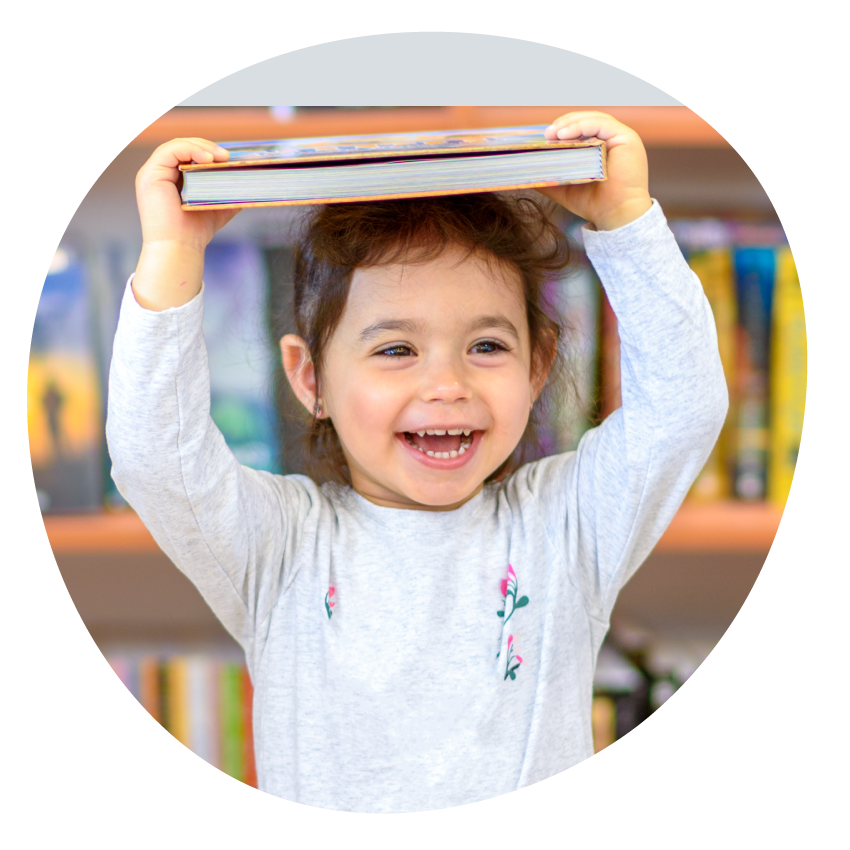 Email Image: A young child in a library smiles while holding a book on their head.
