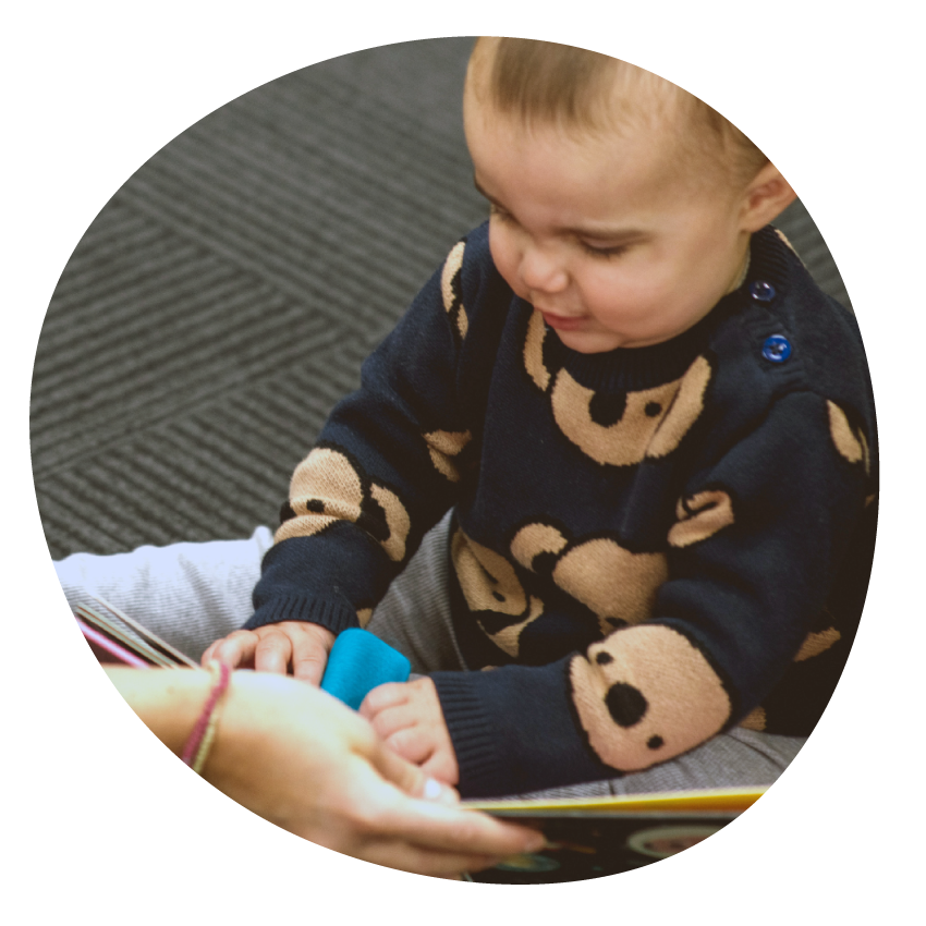 Image: An adult holds a board book for a toddler seated on the floor.