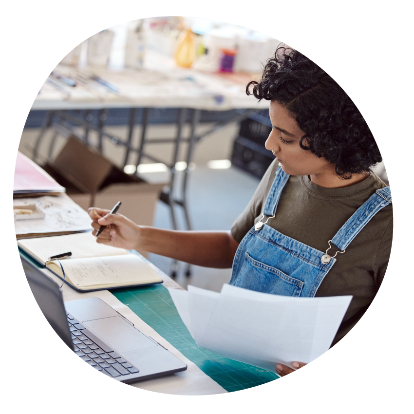 Image: A person sits at a laptop holding sheets of paper and writing in a notebook. 