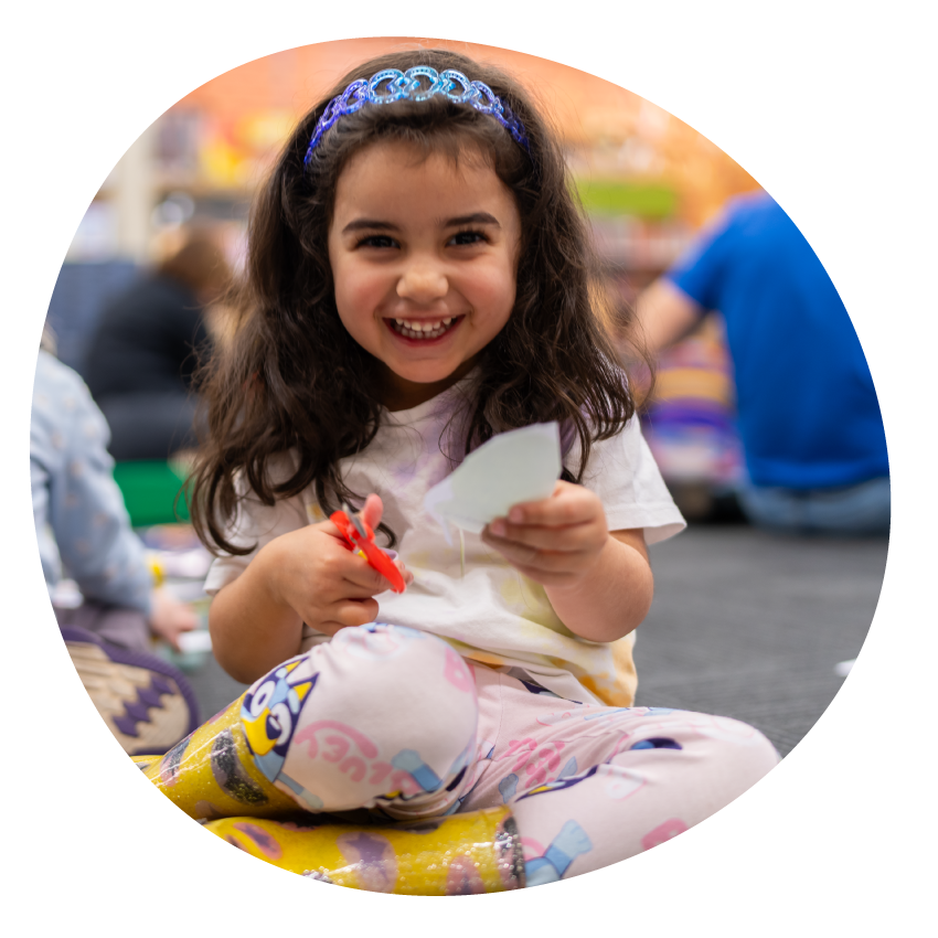 Email Image: A smiling young child sits on the floor at the library doing craft.