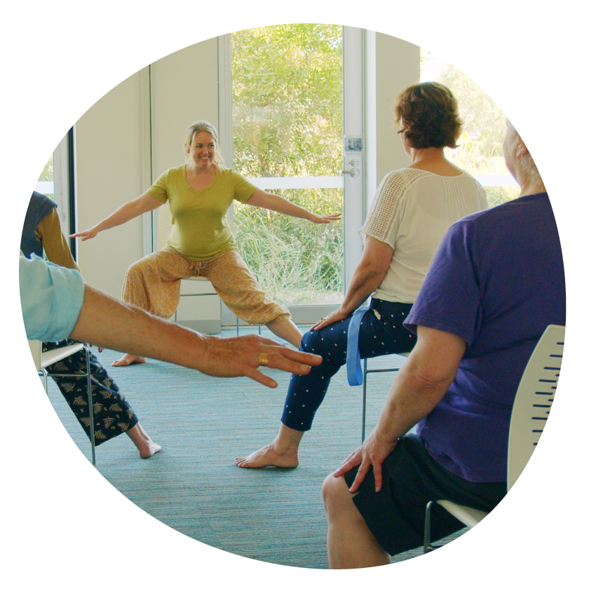 Image: Adults in various seated yoga poses face a yoga instructor conducting a class.