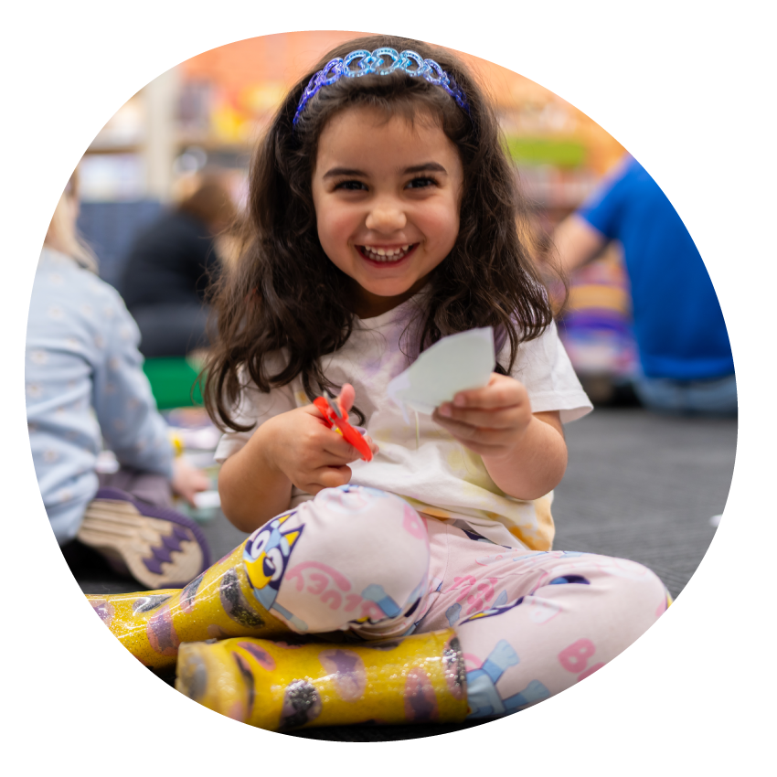 Image: A smiling young child holding craft paper and scissors, with blurry children and adults in the background.