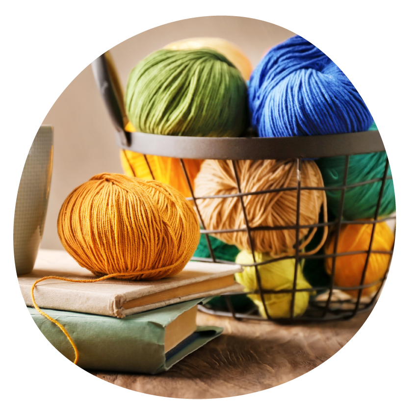 A basket containing coloured balls of wool, two books, a coffee mug and another ball of wool on a table.