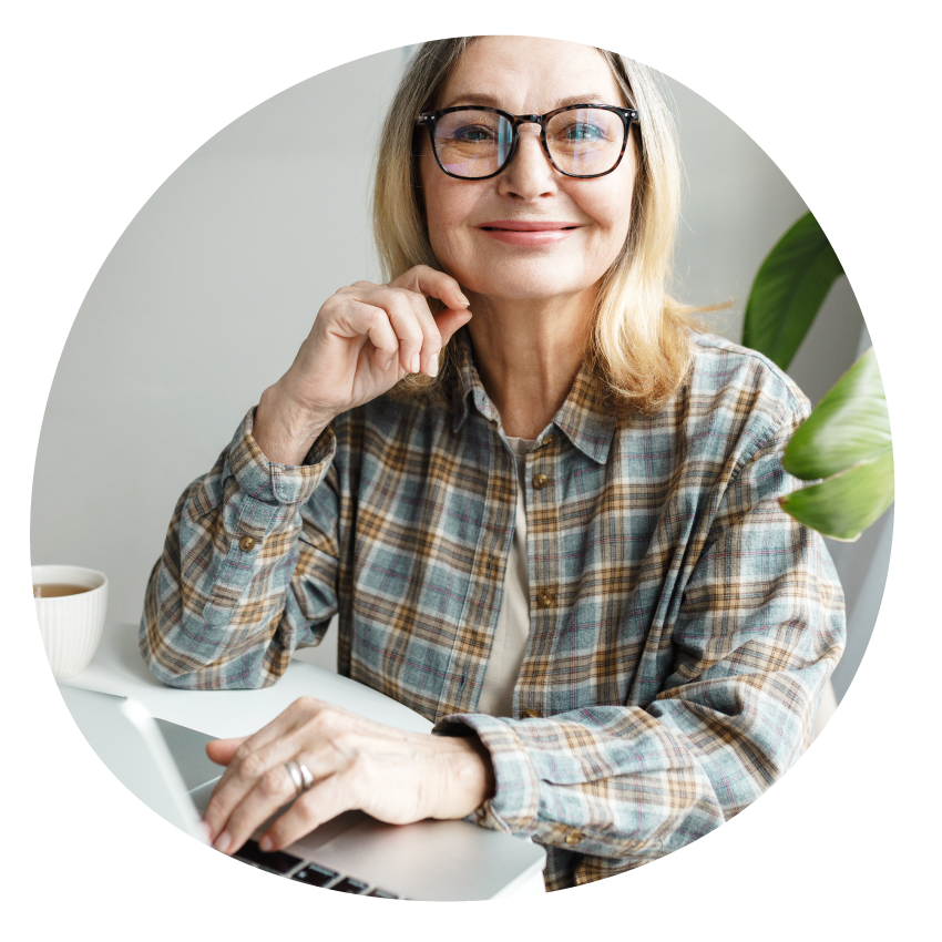  A smiling person in glasses sits at a laptop with a cup of tea at their elbow.