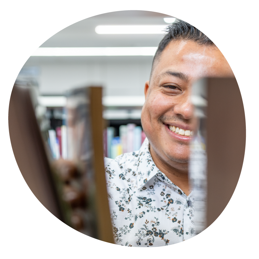 Image: A smiling person glimpsed through books on a library shelf reaches for a book.