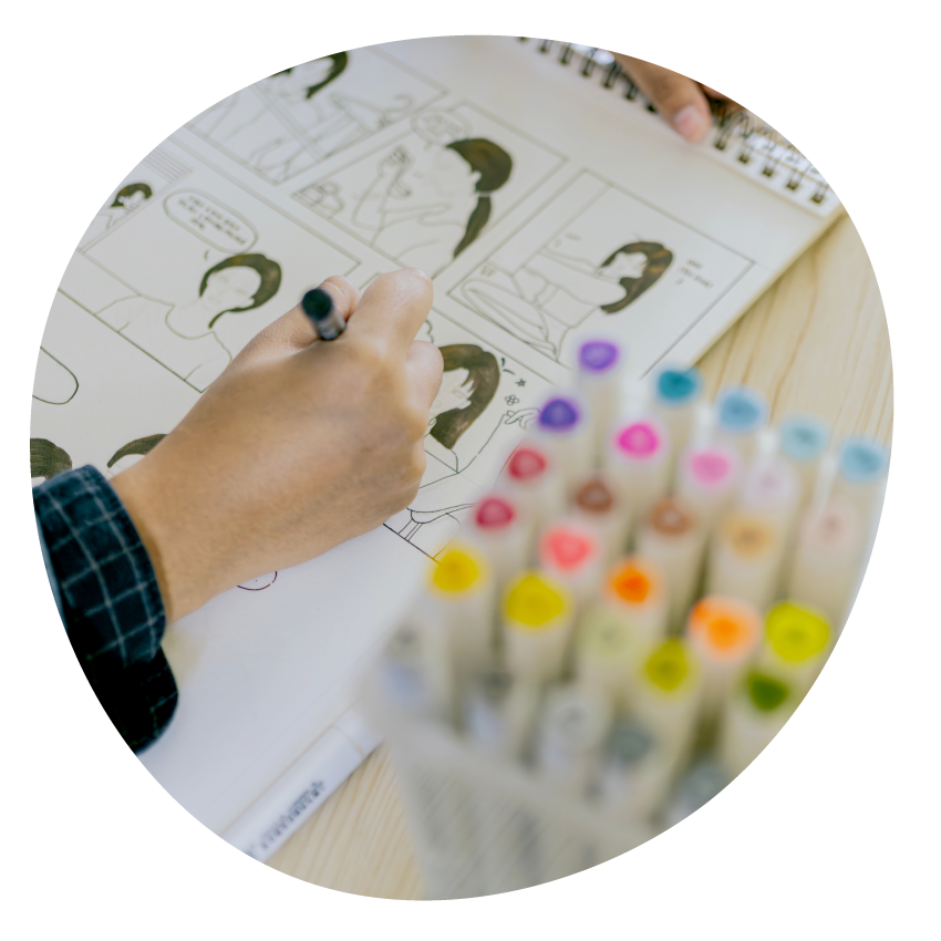 Image: Close-up of a hand drawing a comic strip. Coloured textas sit next to their notebook.