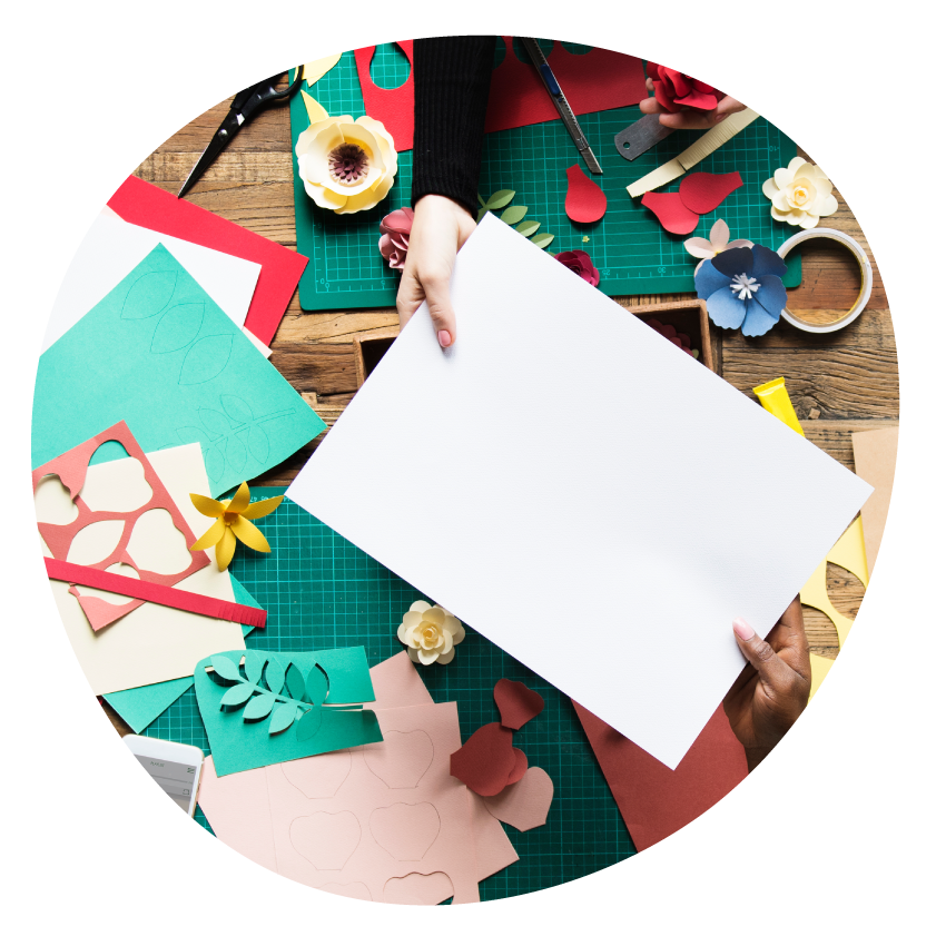 Several pairs of hands doing paper craft at a table covered with cutting boards, paper flowers and foliage, and other craft supplies.