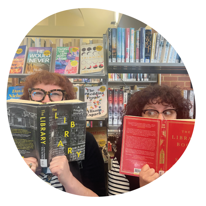 Image: Two library staff members playfully peeking over the top of the open books held in front of their faces.