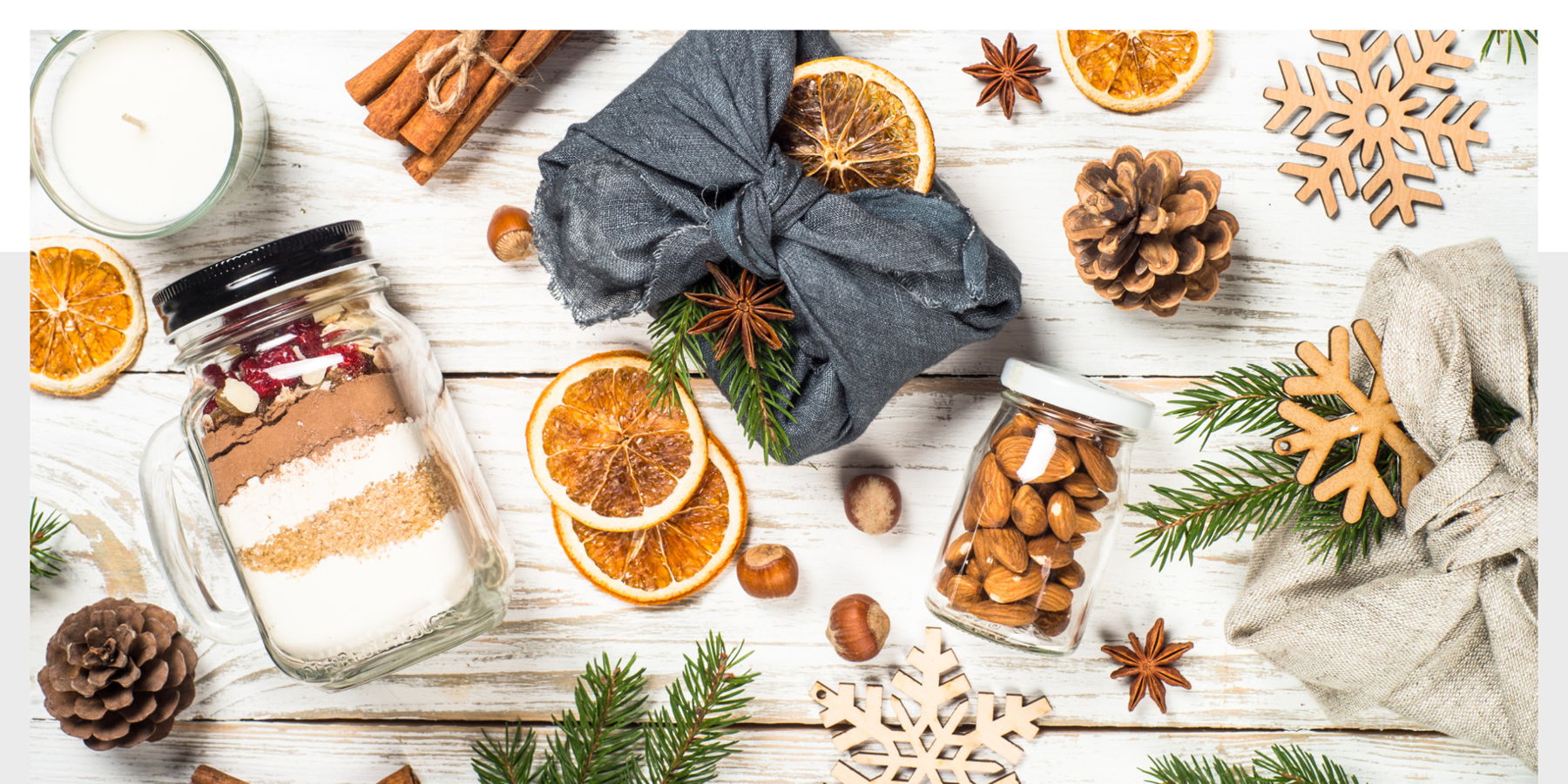 Image: A wooden tabletop scattered with fabric wrapped gifts, wooden snowflakes, cloves, pinecones, dried oranges and jars of food.