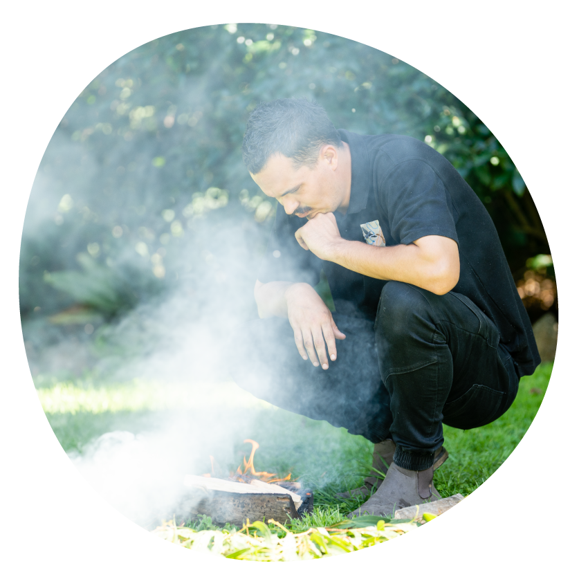 Image: Aboriginal educator Thane Garvey-Gannaway crouched in a grassy area conducting a Smoking Ceremony.