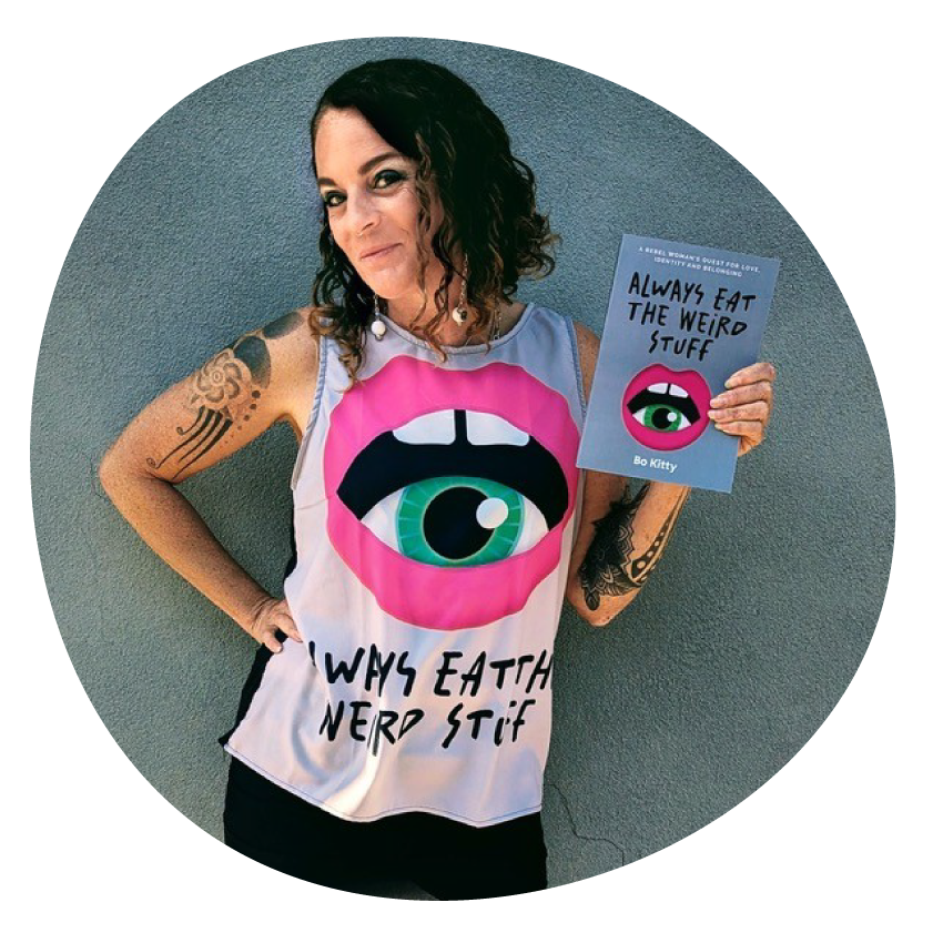 Image: Author Bo Kitty smiles while holding a copy of her book Always Eat the Weird Stuff and wearing a singlet promoting the book.