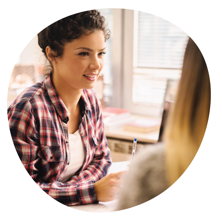 Email Image: A young adult smiling and holding a pen is seated at table looking at another person.