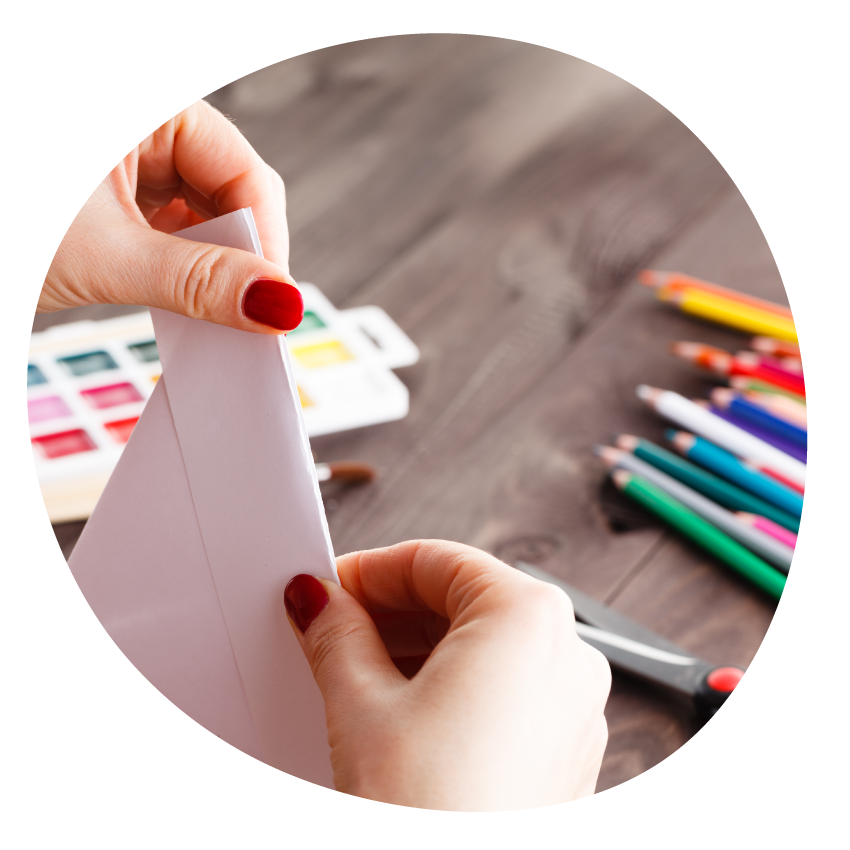 Image: A close-up of two hands folding a piece of paper at a table covered with coloured pencils, a pair of scissors and water colour palette.