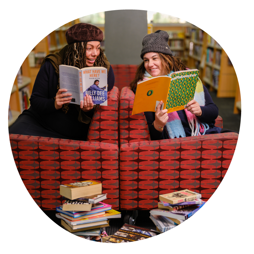 Email Image: Two smiling people wearing beanies are seated back-to-back in library chairs smiling and looking at the books they are each holding.