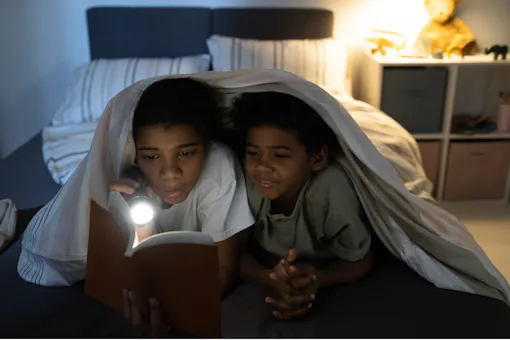 Two children reading under a blanket with a flashlight