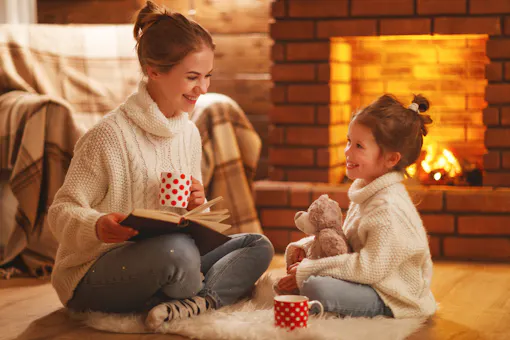 Mother and child reading together by the fireplace enjoying hot chocolate and the child is hugging a teddy bear