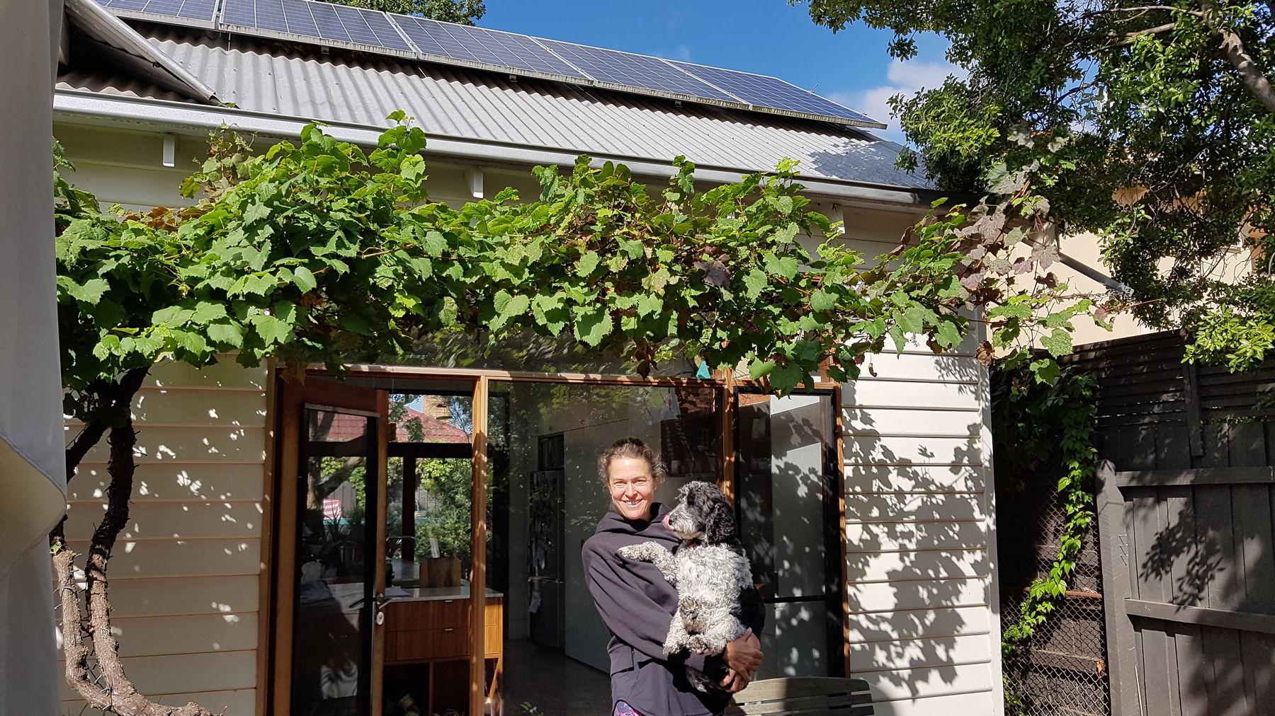 Woman holding dog standing in front of house with solar panels under a grape vine