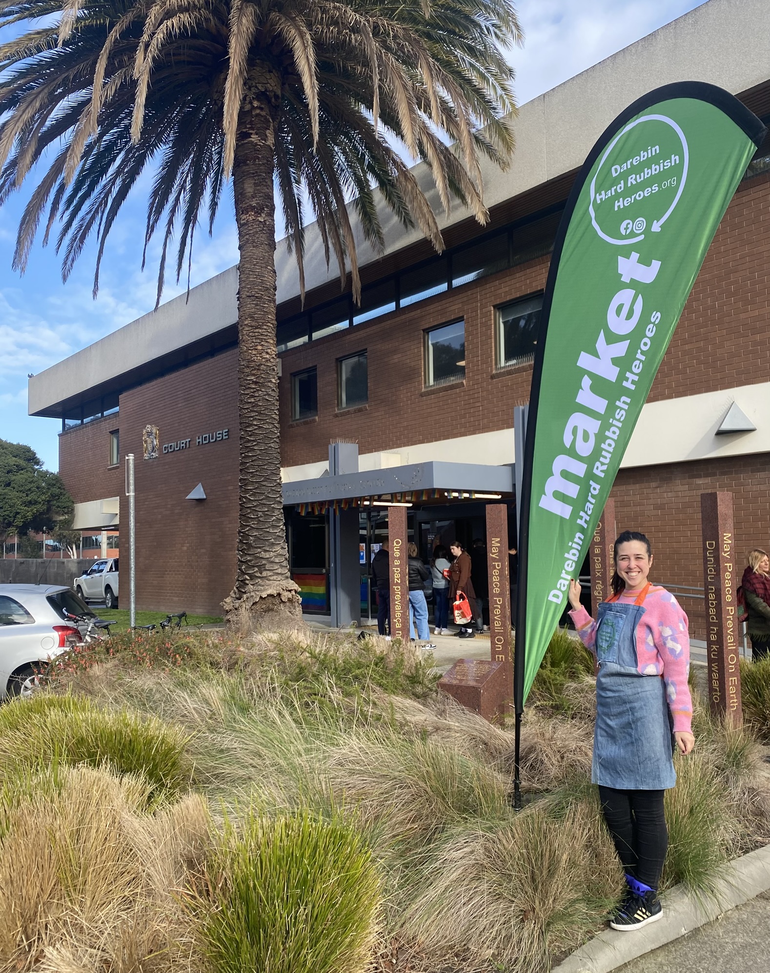 Picture of a person standing next to a banner outside a building. The banner reads 'Market Darebin Hard Rubbish Heroes'
