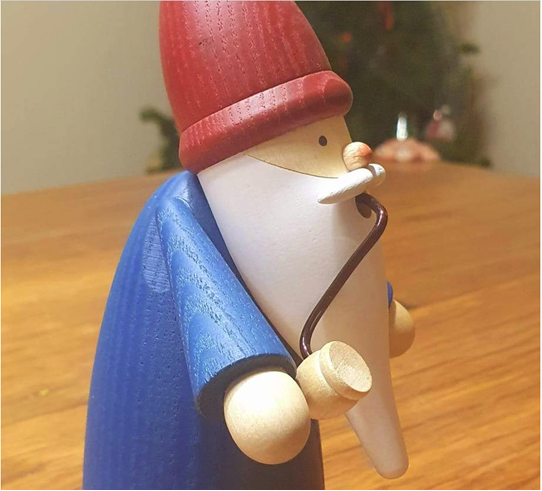 Photo of a wooden figure in a red hat and blue clothes with a long white beard smoking a pipe with a Christmas tree in the background