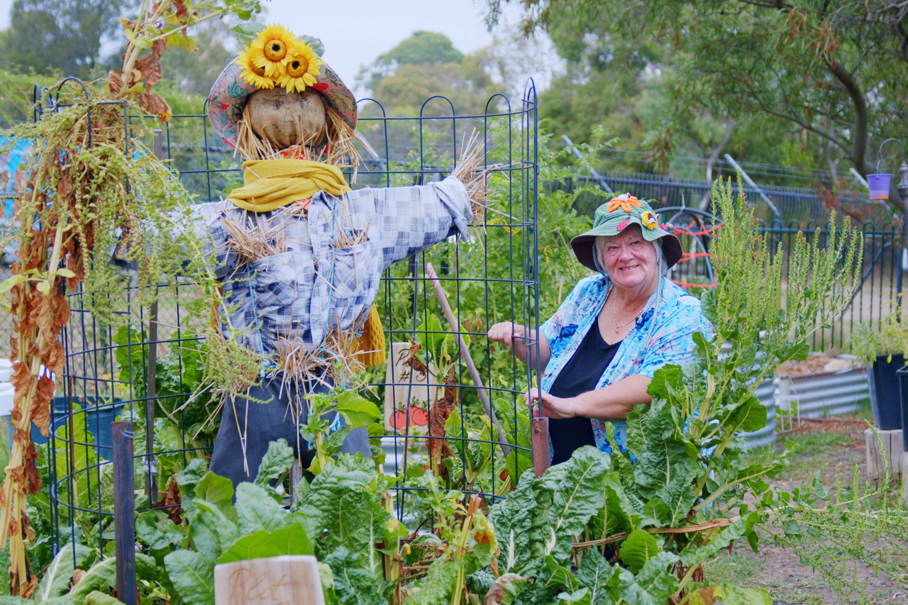 Woman in a garden wearing a floppy hat standing next to a big scarecrow with sunflowers attached to its head