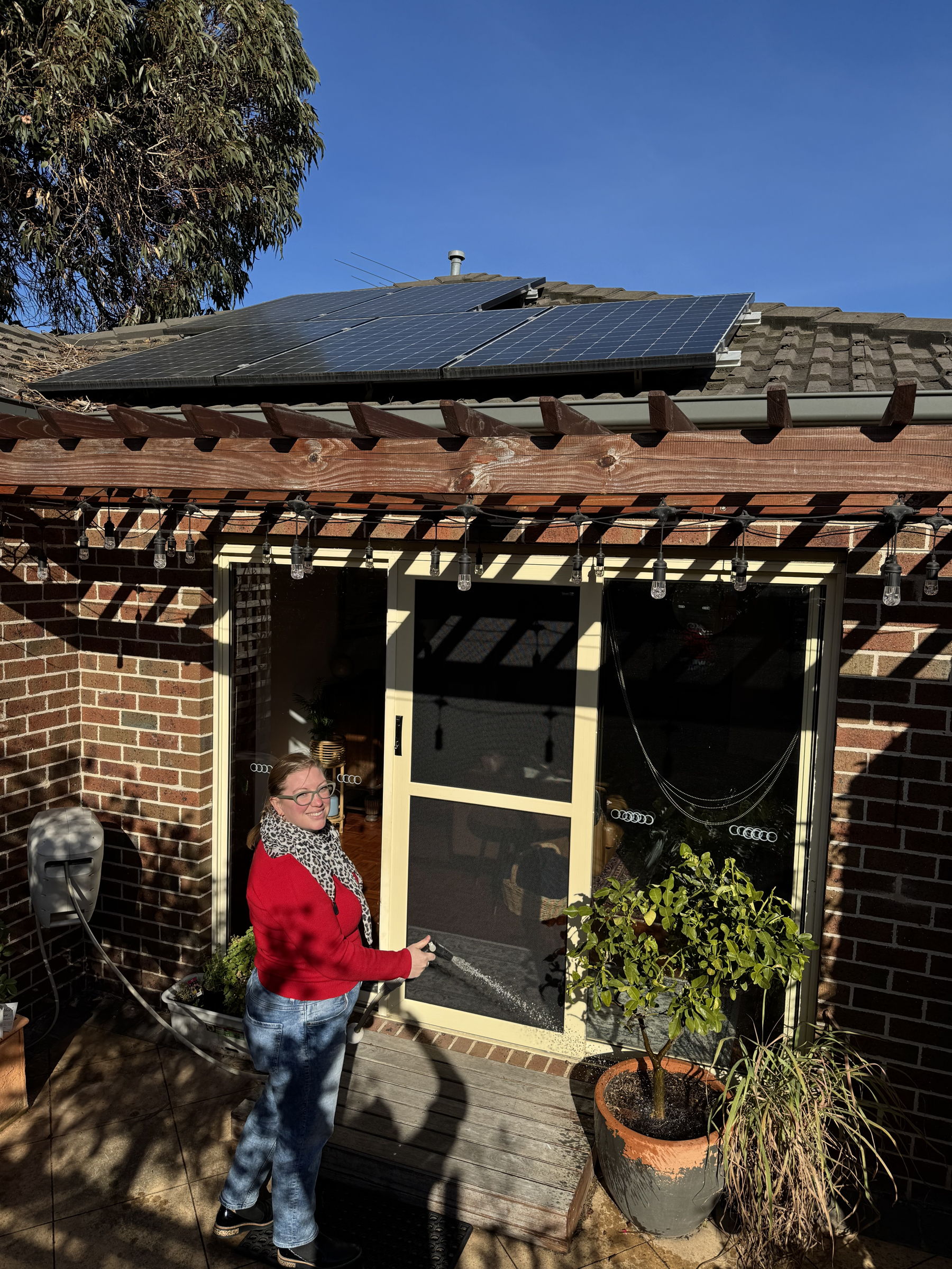 Emily watering her garden, her solar panels are visible on her roof in the background