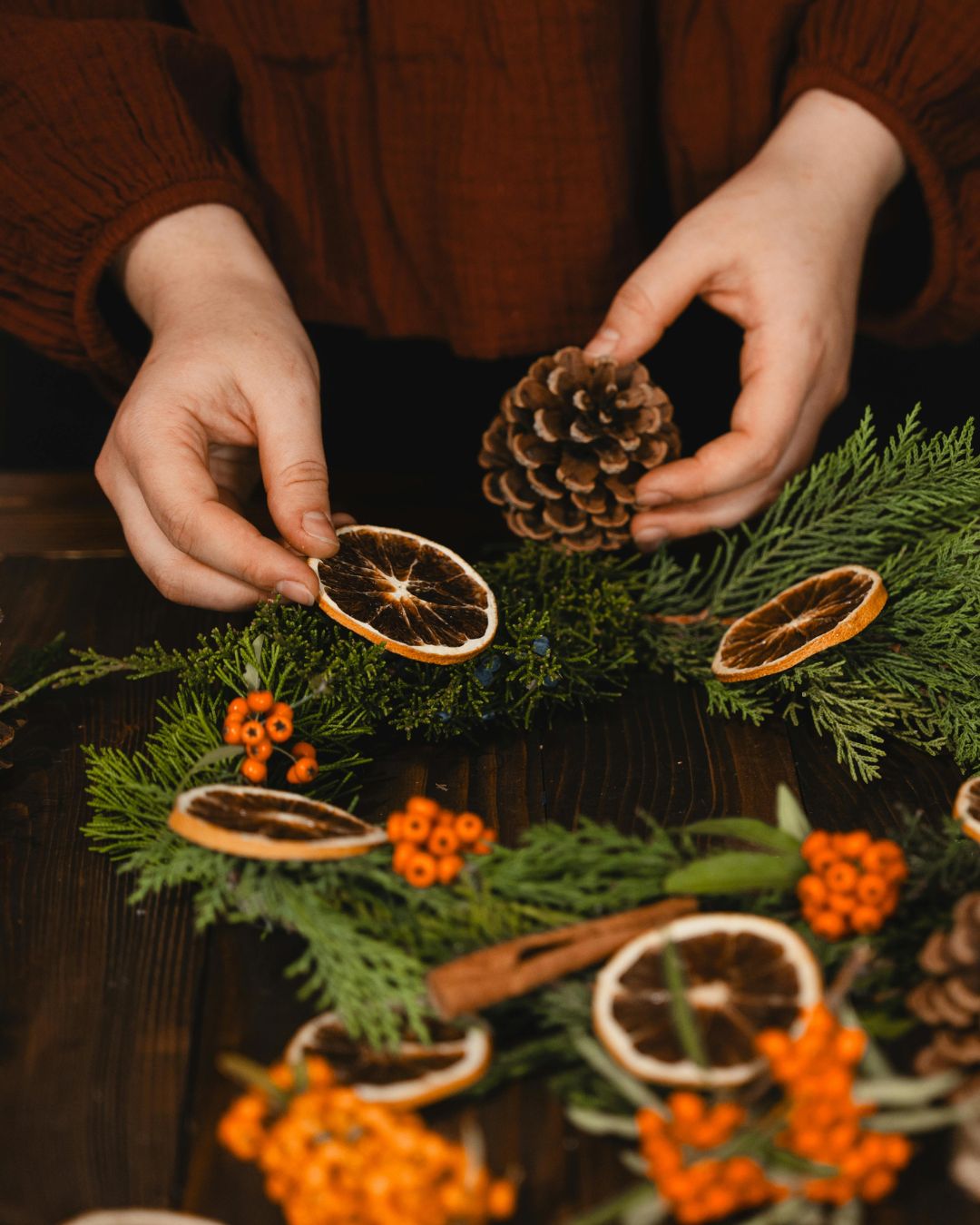Person's hands putting together a festive wreath with dried orange slices, cinnamon and pine cones