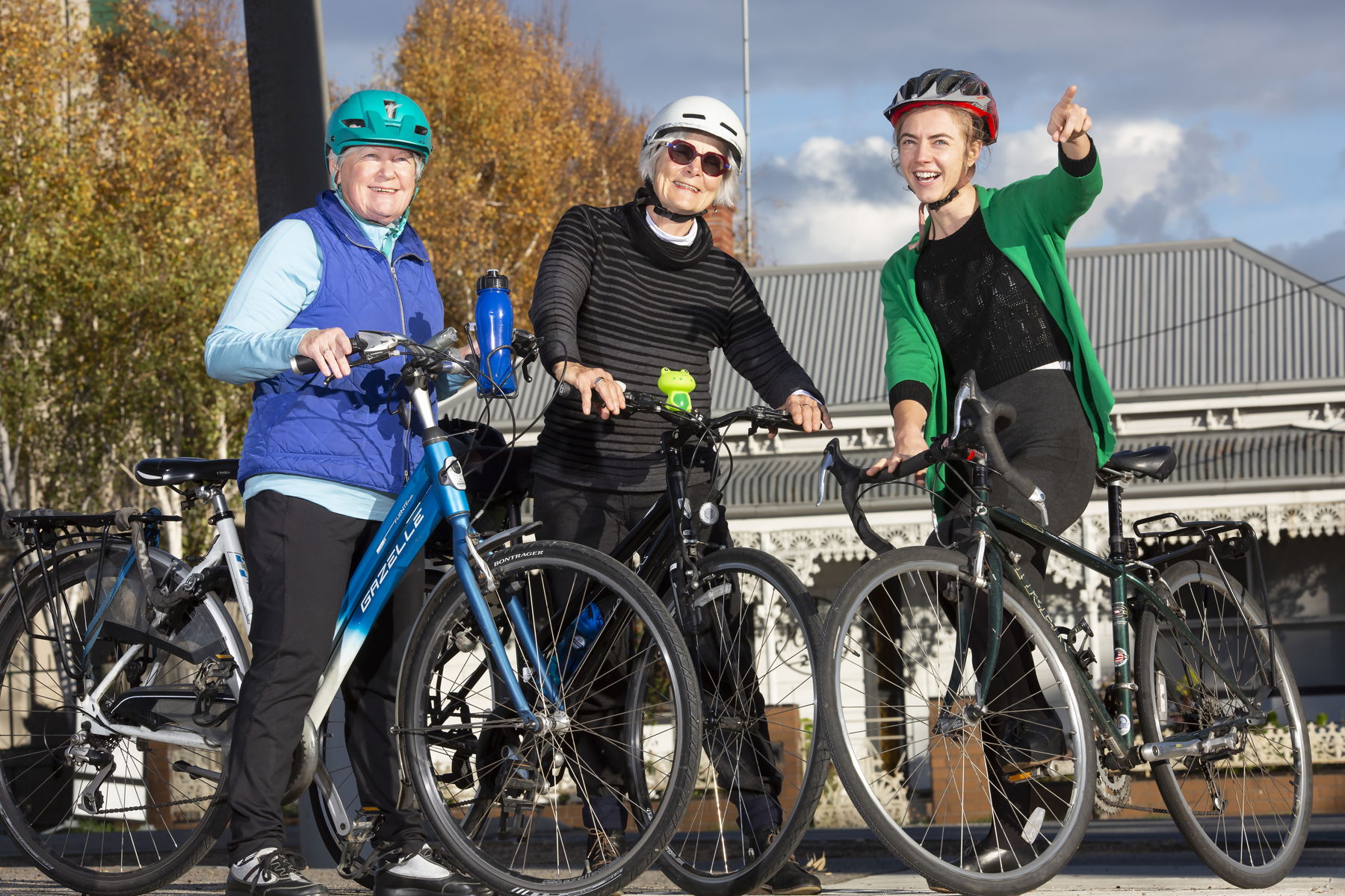 Three women standing with their bikes on the street out the front of a white house.