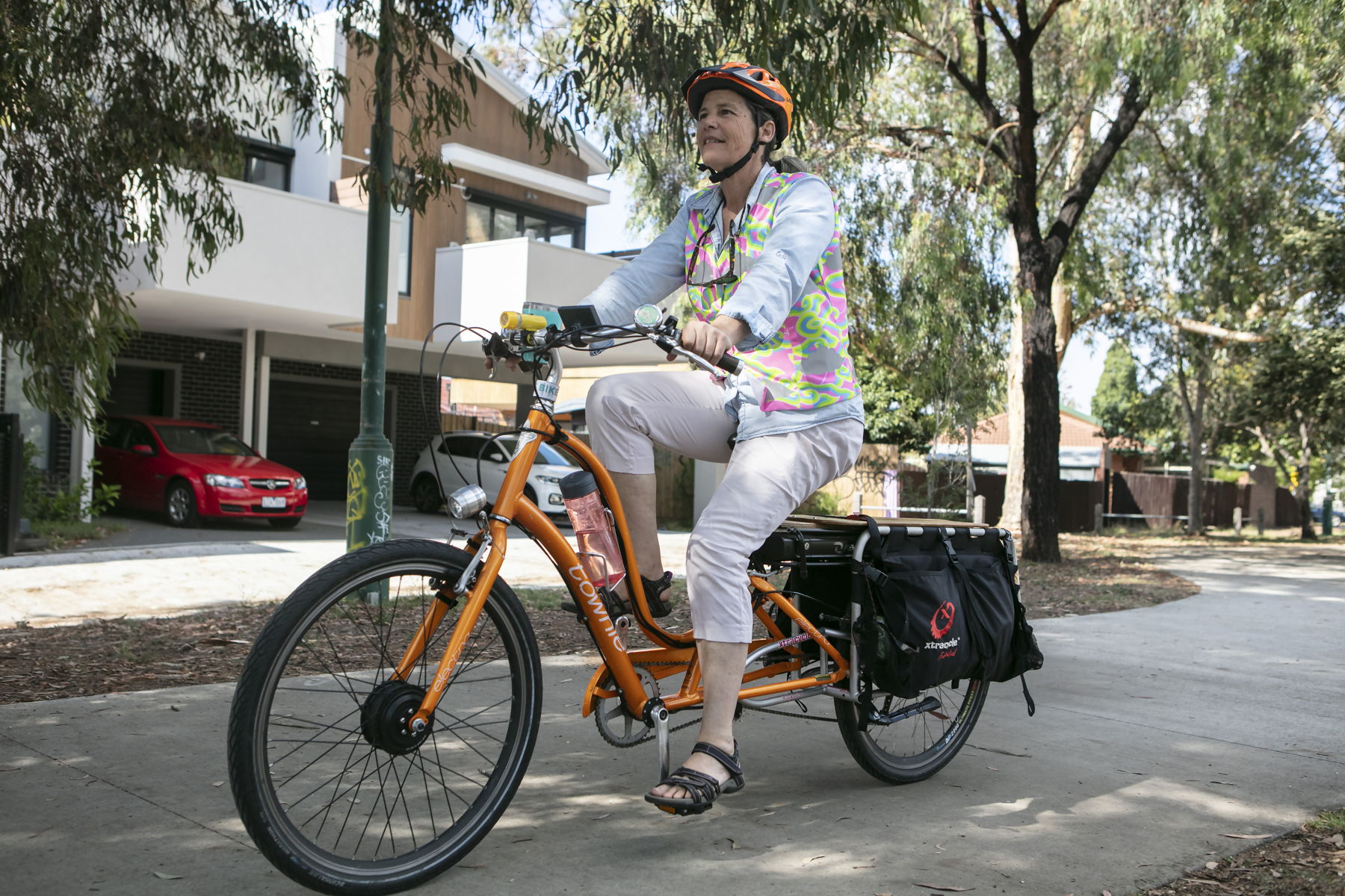 Woman riding a bright orange e-cargo bike with panniers down a shady path