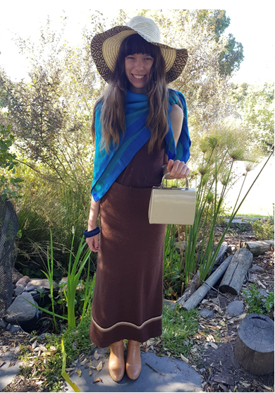 Woman wearing a long brown dress with a bright blue scarf tied around her neck and a floppy hat on her head, holding a little handbag. She's standing in a garden.