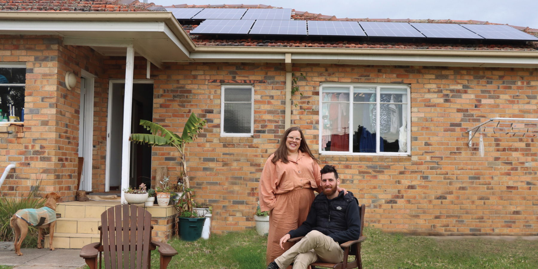 Photo of Steph Rich and her partner out the front of their home - you can see solar panels on their roof in the background.