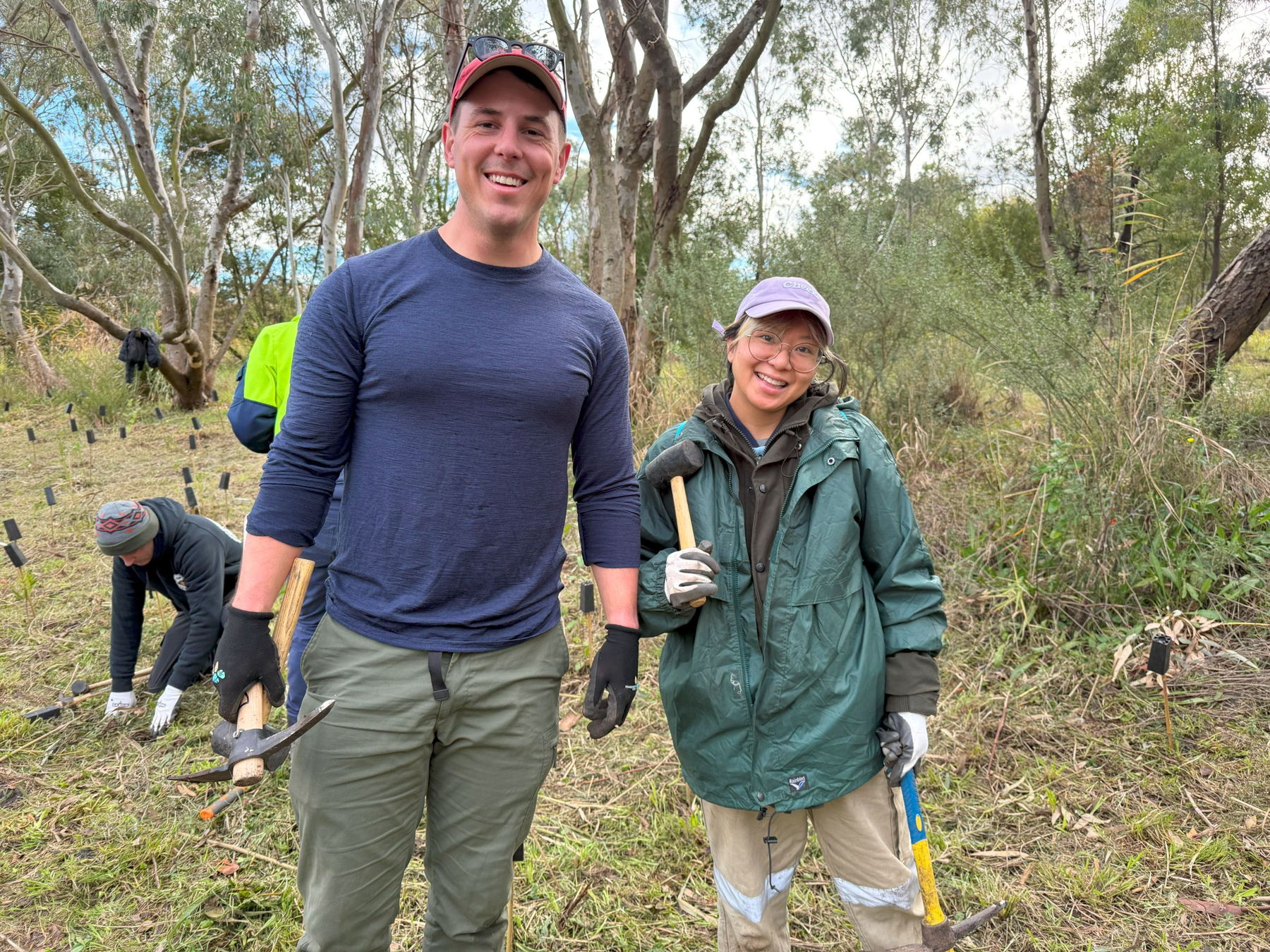 two people smiling at the camera with gloves on ready to plant in front of trees
