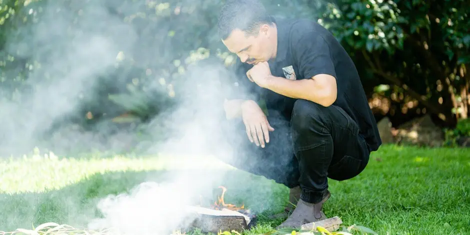 Aboriginal educator Thane Garvey-Gannaway crouched in a grassy area conducting a Smoking Ceremony.