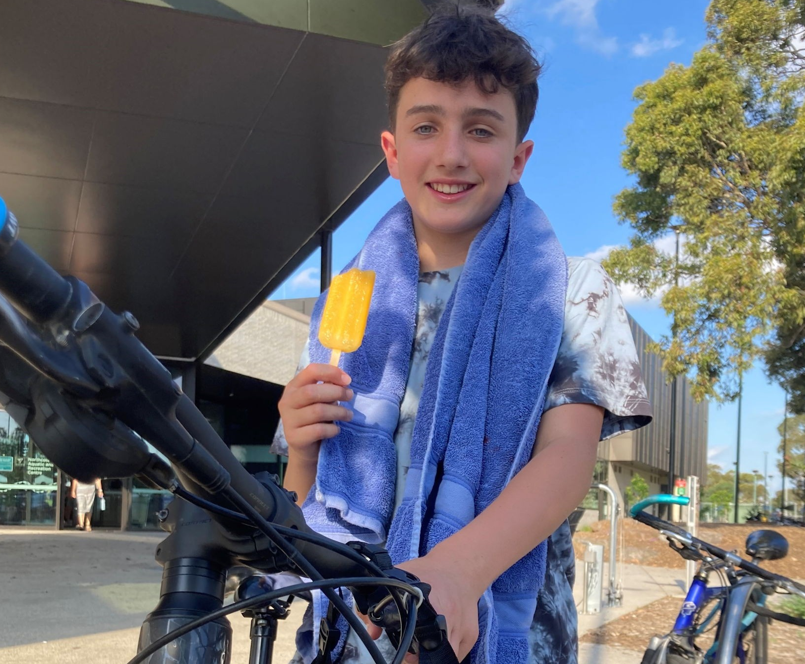 Person on a bike holding an icy-pole standing outside Northcote Pool with a towel around their neck and holding their bike